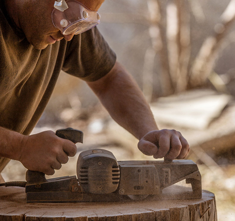 Carpenter working with electric planer on wooden stump in the open air, wearing goggles.